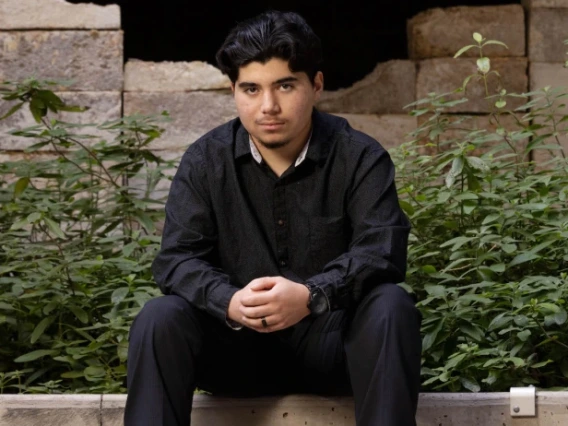 Marcelino Perez, dressed in black pants and shirt, sits on concrete wall facing the camera.