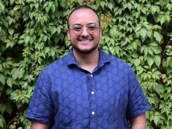 Key Sanchez wearing glasses with a blue button up shirt in front of foliage. 