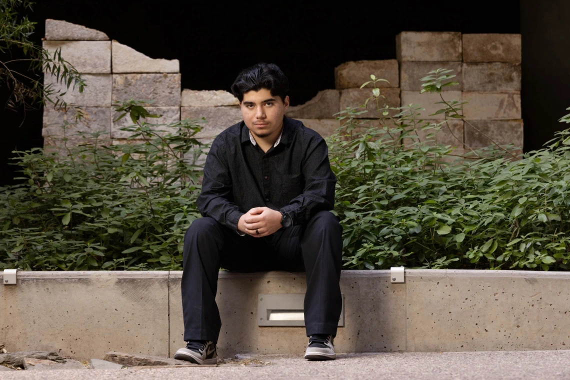 Marcelino Perez, dressed in black pants and shirt, sits on concrete wall facing the camera.