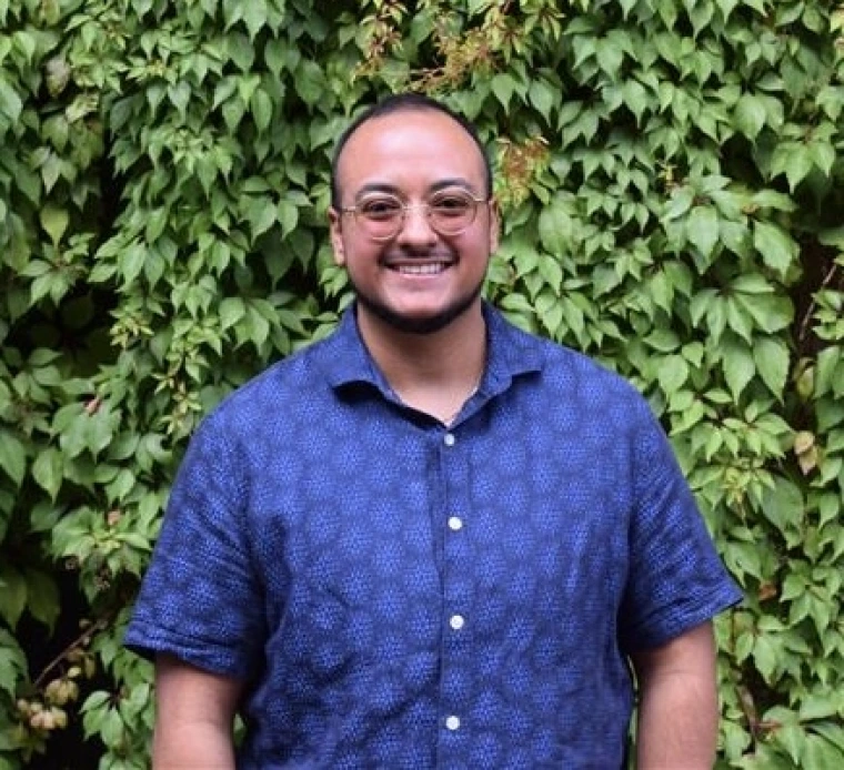 Key Sanchez wearing glasses with a blue button up shirt in front of foliage. 