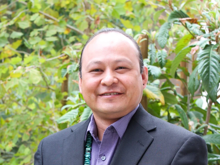 Derwin Begay's Portrait. He is posing in front of green trees, smiling and wearing a purple dress shirt and a black suit coat. 