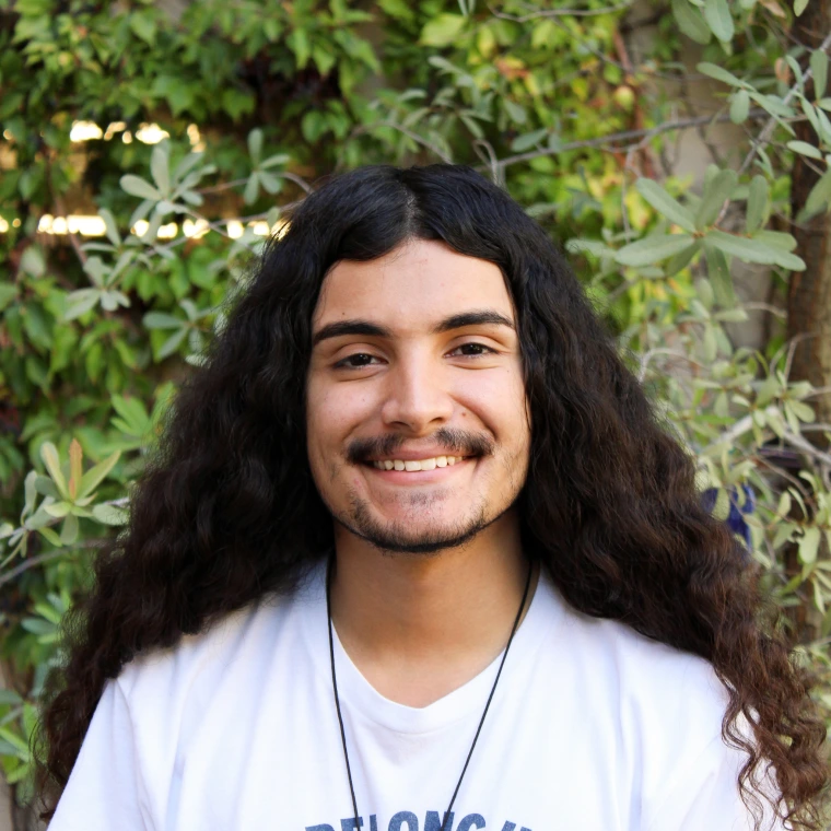 Roman Orozco, wearing a white shirt, smiling to the camera in front of a green foliage background.