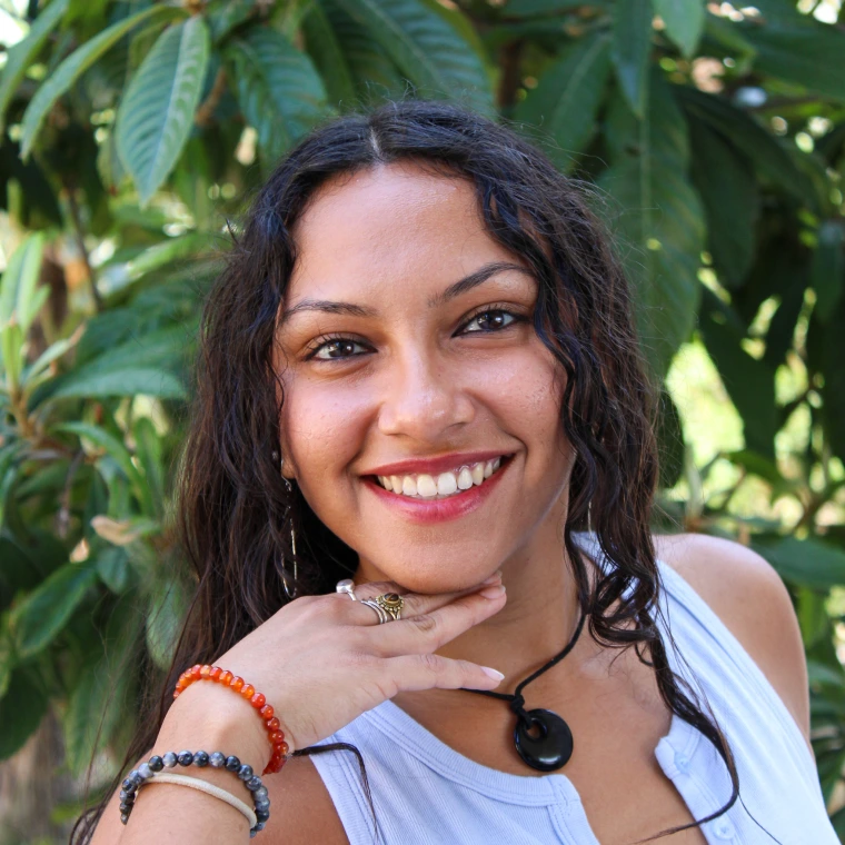 Keily Cruz, wearing a light gray tank top. Smiling posing to the camera with her hand underneath her chin and in front of a green foliage background.