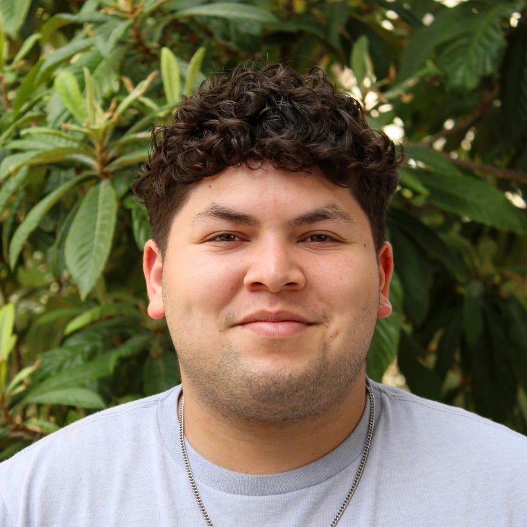 Enrique Rojo, wearing a light great t-shirt. Smiling posing to the camera with a green foliage background.