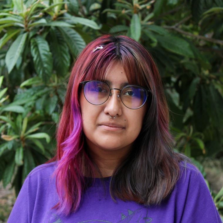 Suni Salas, wearing a purple t-shirt. Smiling posing to the camera with a green foliage background.