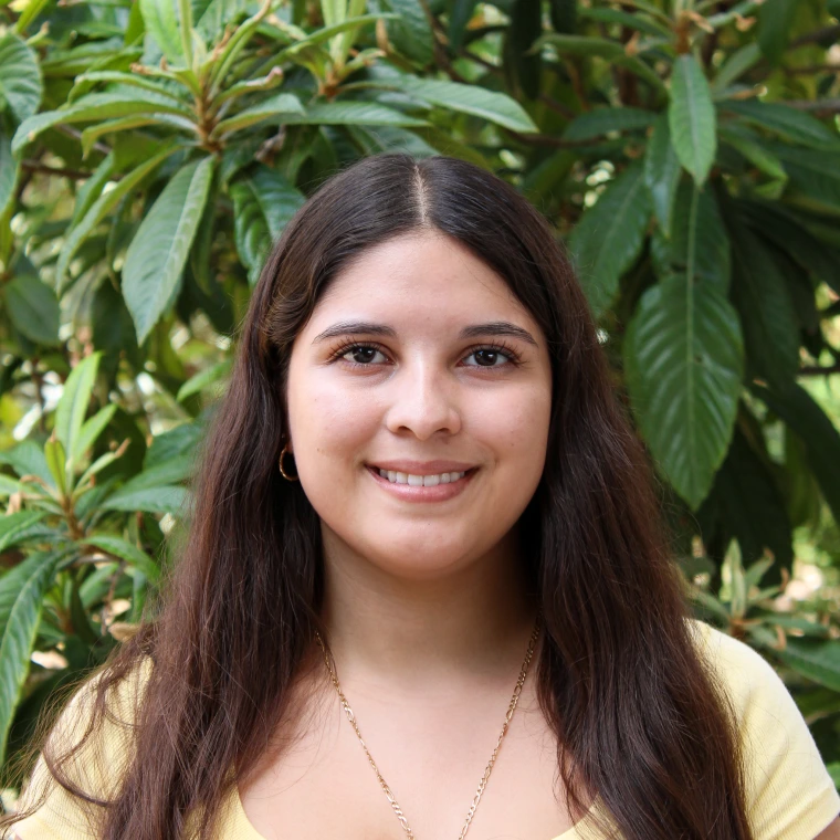 Aracely Cazares, wearing a yellow shirt. Smiling posing to the camera with a green foliage background.
