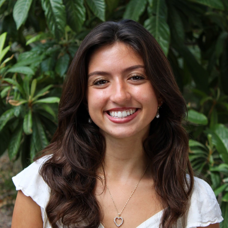 Lucy Stage, wearing a white blouse. Smiling posing to the camera with a green foliage background.