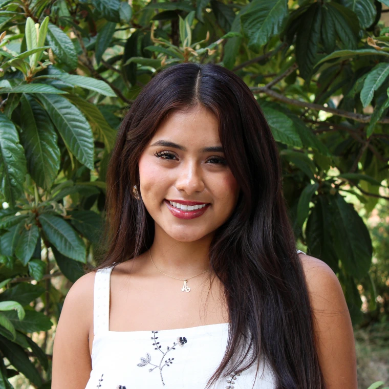 Alexa Garcia, wearing a white blouse. Smiling posing to the camera with a green foliage background.