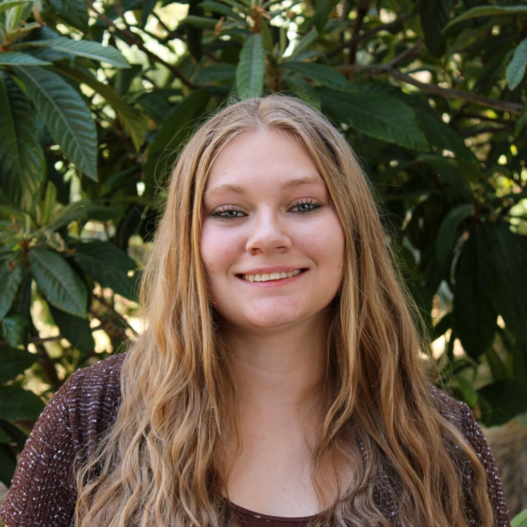 Serenity Matanza, wearing a brown blouse, smiling to the camera in front of a green foliage background.