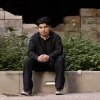 Marcelino Perez, dressed in black pants and shirt, sits on concrete wall facing the camera.